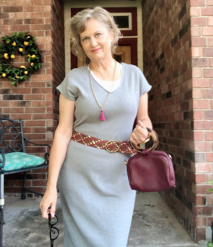 A woman over 60 with a short wavy dark blonde and silver hairstyle swept to the side while posing outdoors in a gray dress.
