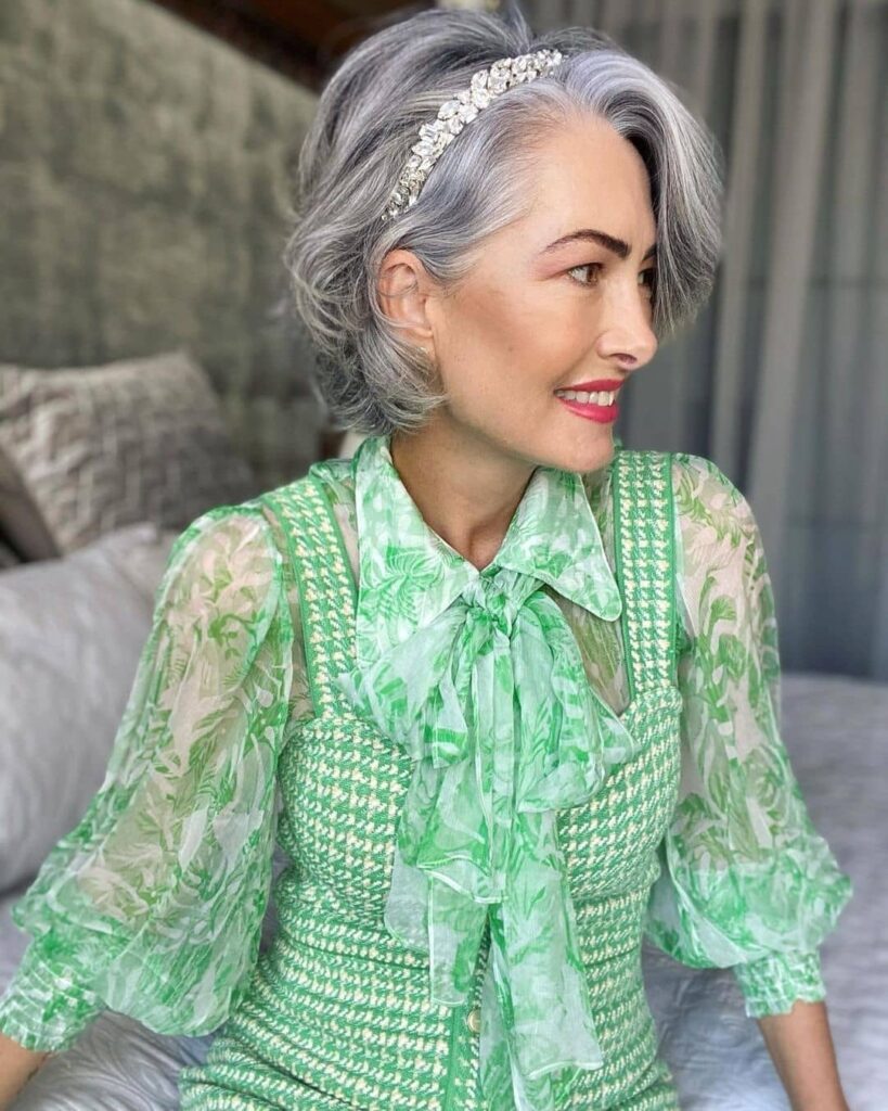 A woman with a short layered wavy silver bob hairstyle wearing a sparkly jeweled headband and a green floral blouse.