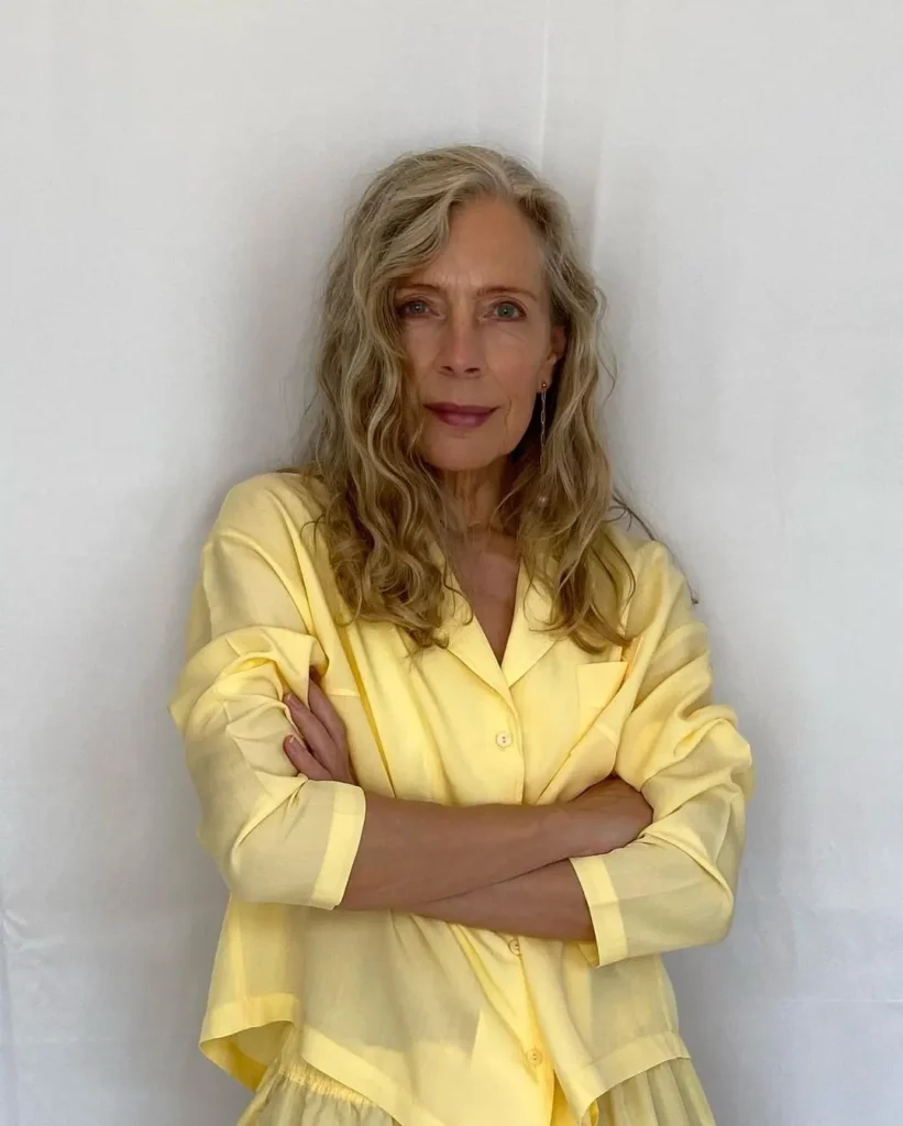 A woman over 60 with long wavy ash blonde hair and a side part wearing a yellow button down shirt.