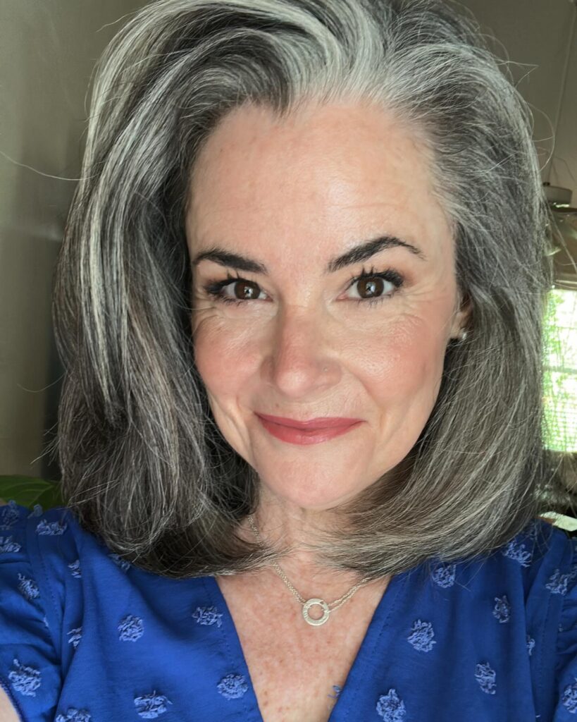 A smiling woman over 60 with a voluminous shoulder-length lob haircut in a natural salt-and-pepper gray color.