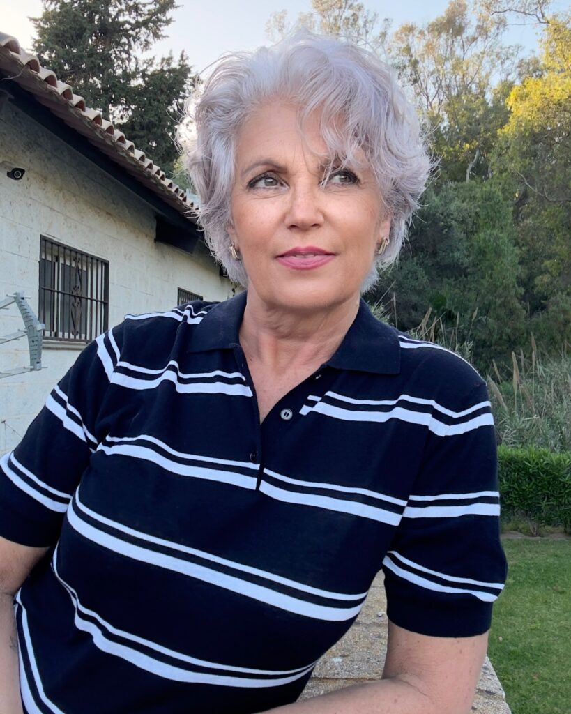 A chic woman over 60 posing outdoors with a short, voluminous silver crop haircut featuring soft natural curls and dark undertones.