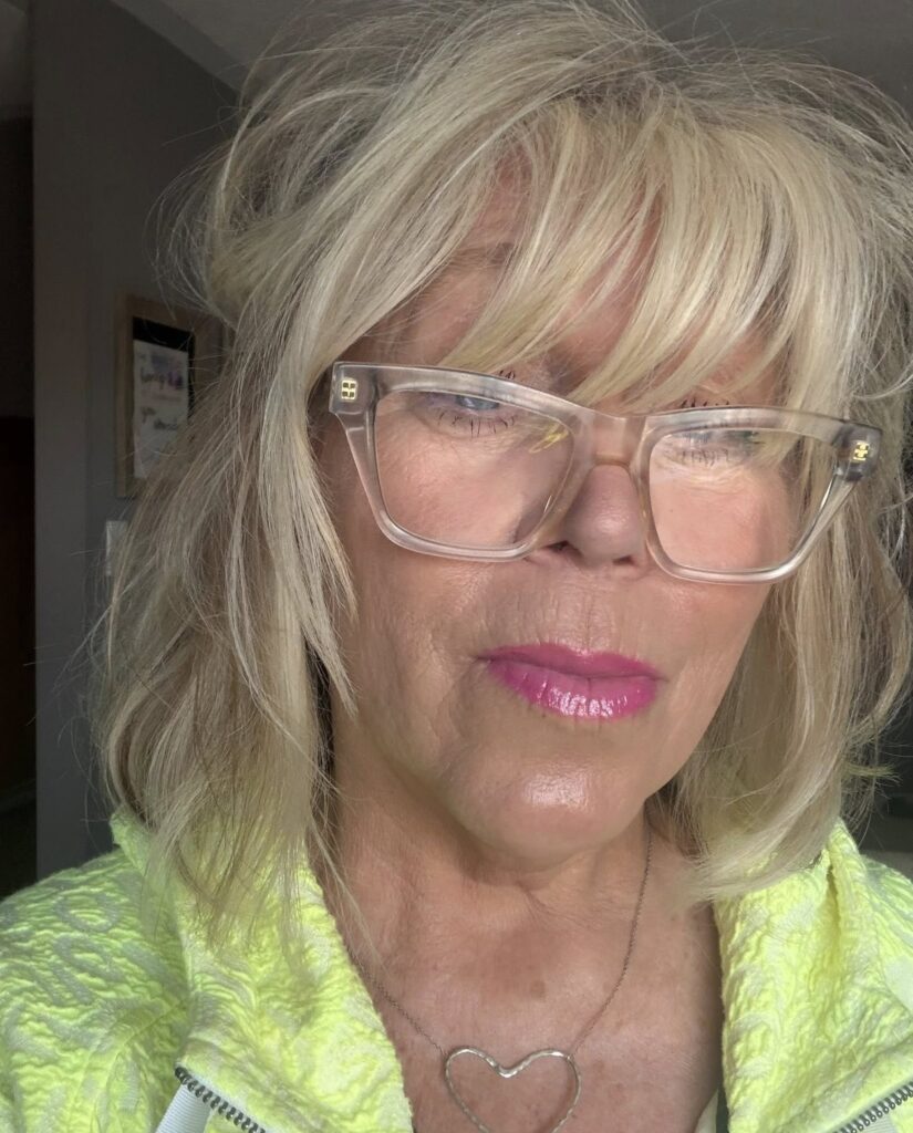 A trendy woman over 60 wearing clear glasses and sporting a textured blonde shaggy lob haircut with thick piecey bangs.