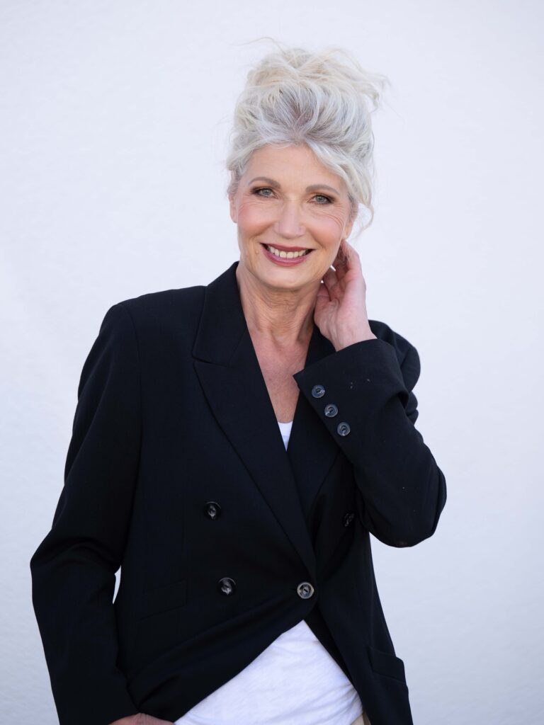 A smiling woman over 60 with long platinum-white hair styled up into a voluminous messy high bun with loose side tendrils.