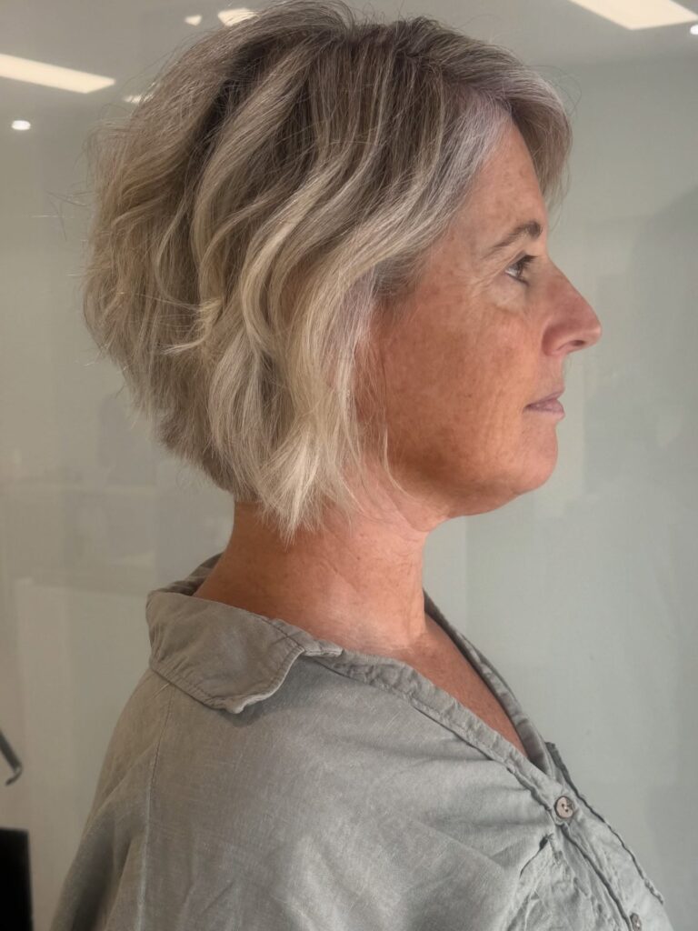 A woman over 60 shown in profile with a textured ash blonde, angled chin-length bob haircut.