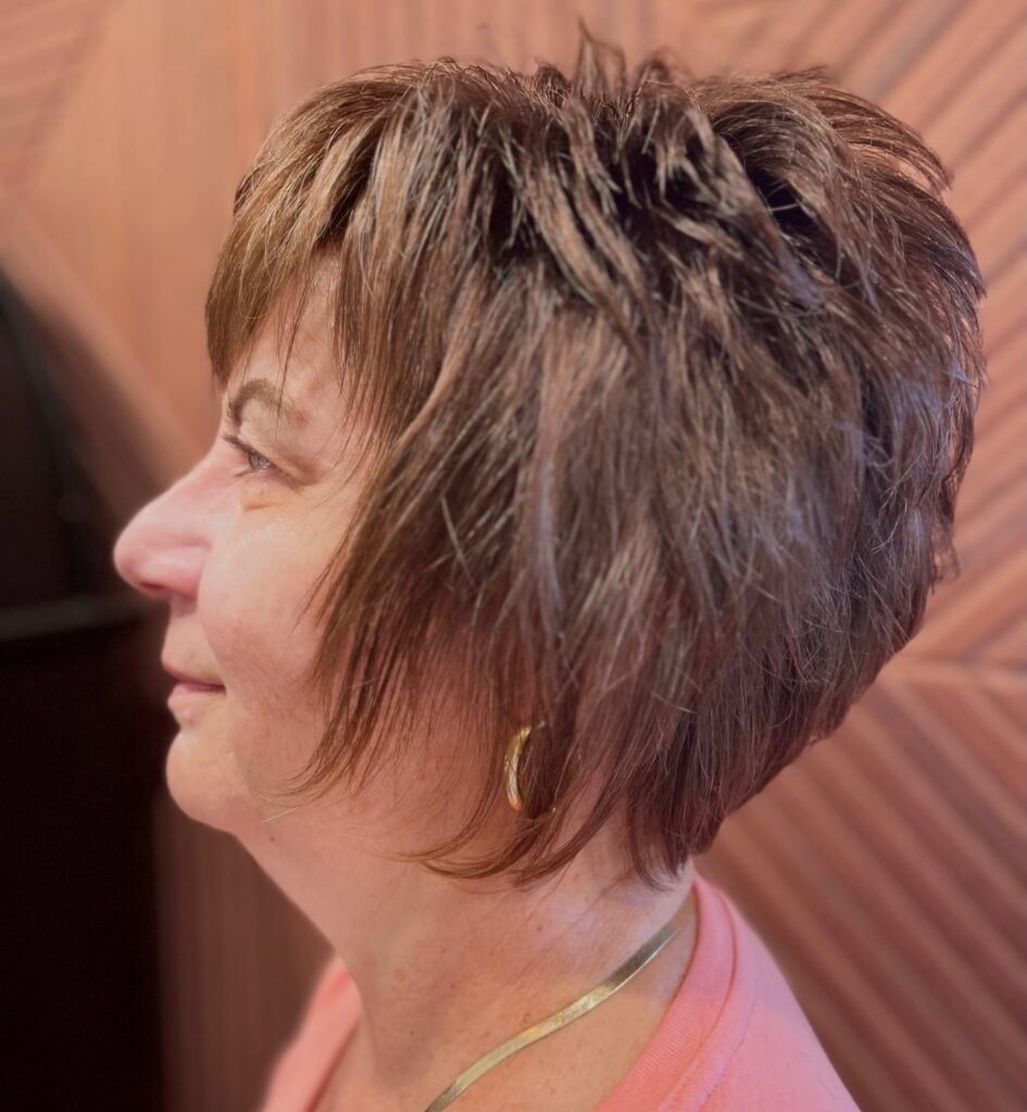 A side profile of a woman over 60 with a choppy caramel brown pixie bob haircut featuring voluminous textured layers and wispy bangs.