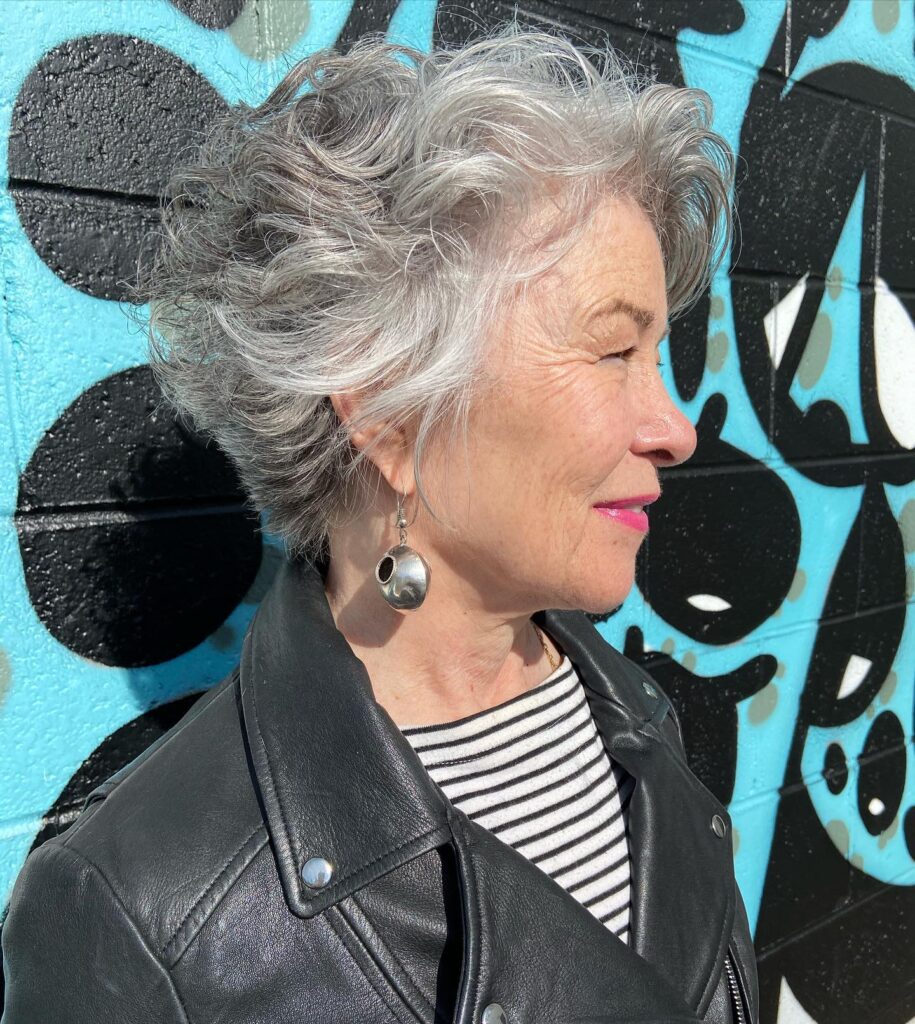 A side profile of a woman over 60 with a voluminous silver and gray textured pixie haircut featuring tousled curls.