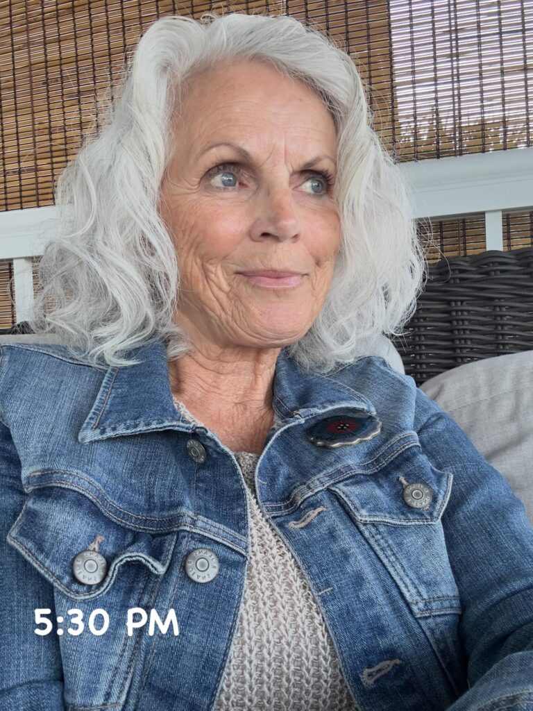 A woman over 60 with shoulder-length wavy silver hair wearing a denim jacket.