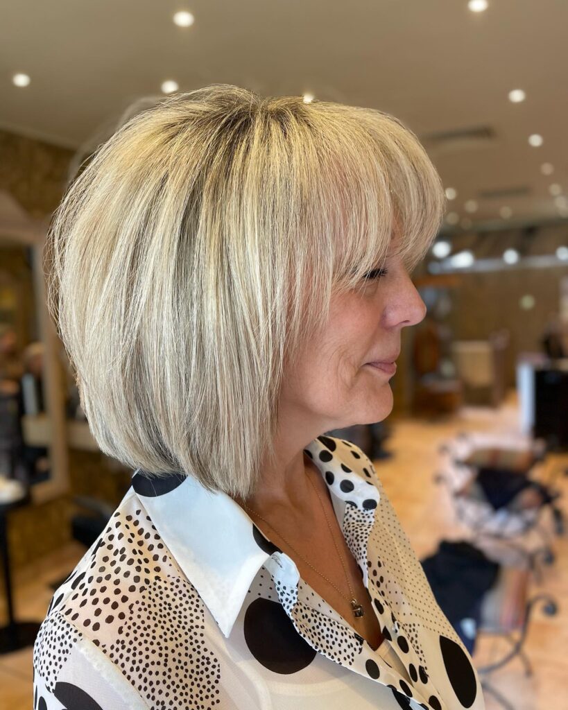 A woman over 60 shown in profile with a sleek, chin-length, bright blonde, rounded bob haircut featuring soft, wispy bangs.