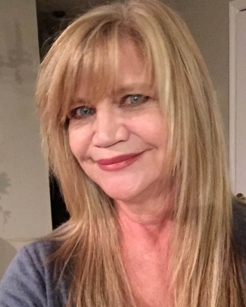 A smiling woman over 60 with long golden blonde straight hair featuring face-framing layers and wispy bangs.