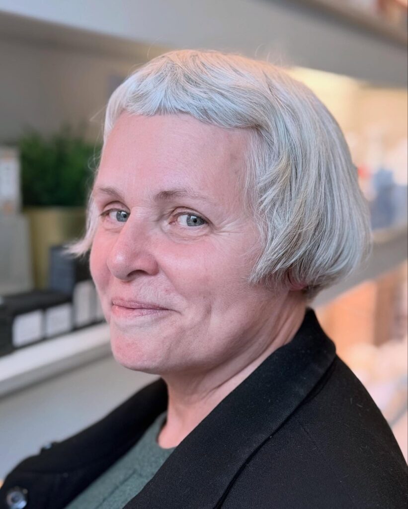 A smiling woman over 60 with a very short, ear-length, straight silver micro-bob haircut featuring a curved micro-fringe.
