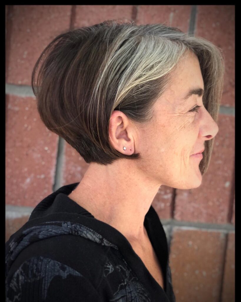A side profile of a woman over 60 sporting an asymmetrical dark brunette wedge bob with a prominent natural silver streak in the front.