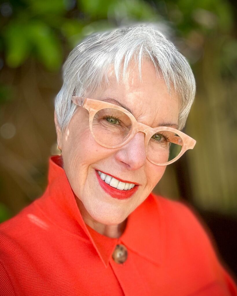 A smiling woman over 60 with a very short, sleek silver pixie cut and micro bangs wearing glasses.