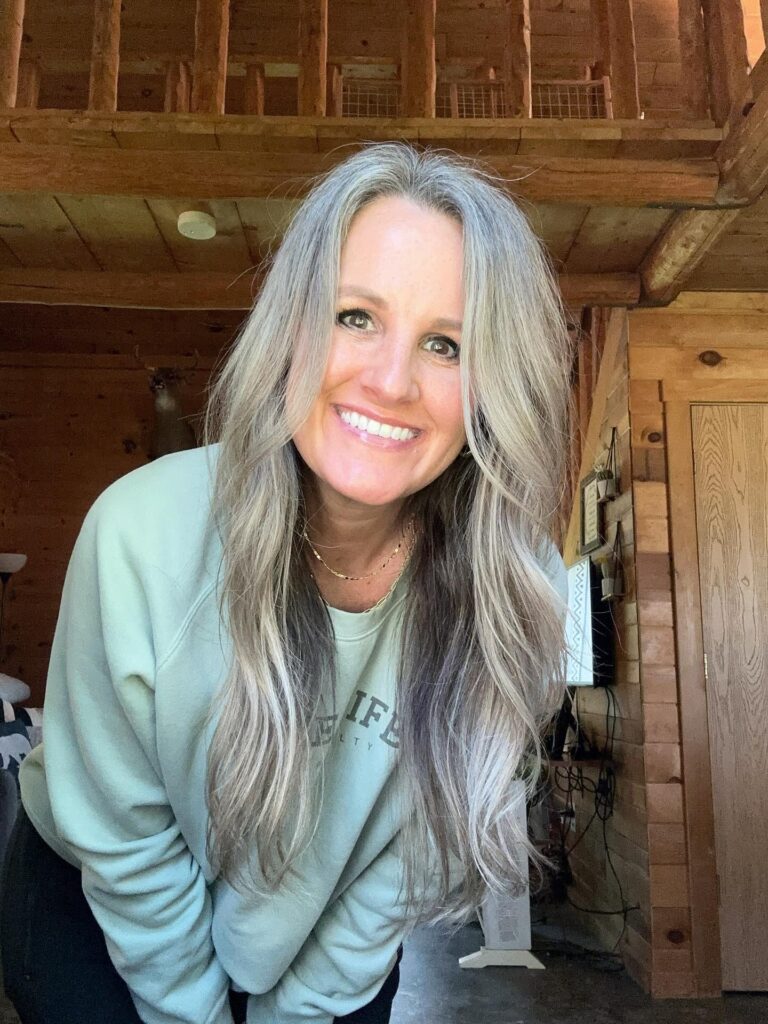 A smiling woman with long layered silver and gray hair wearing a light green sweatshirt in a wooden room.
