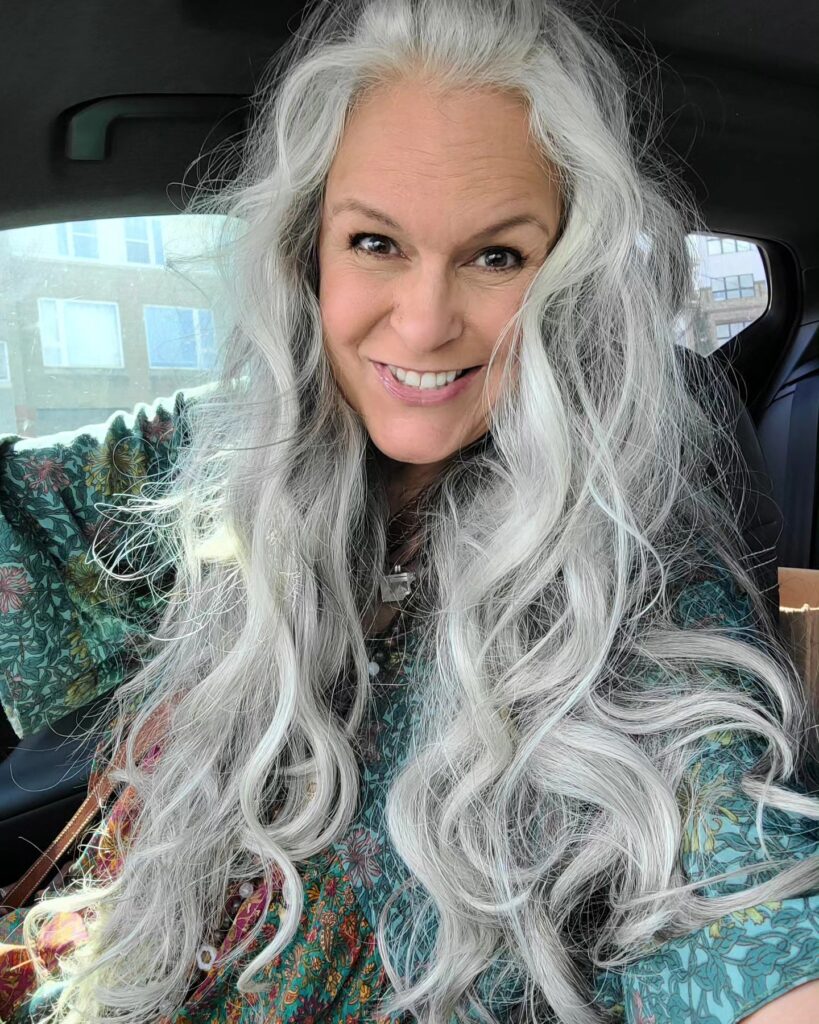 A smiling woman with voluminous, long curly silver hair sits in a car wearing a green patterned top.