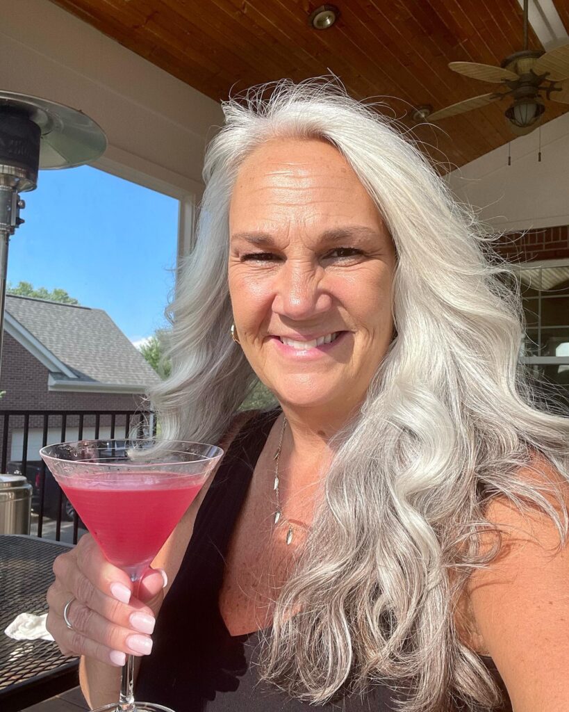 A smiling woman outdoors holding a pink cocktail showcases her voluminous long silver hair styled in soft bouncing waves.