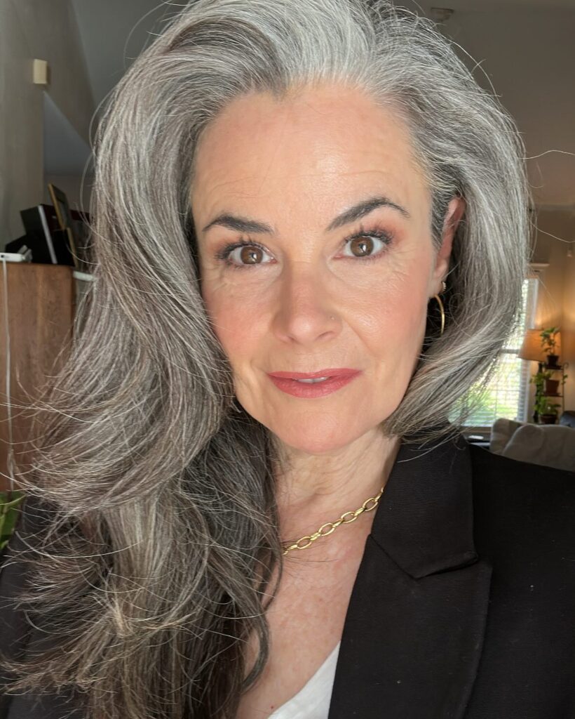 A close up of a woman with voluminous, side-swept salt and pepper gray hair wearing a black blazer and a gold chain necklace.