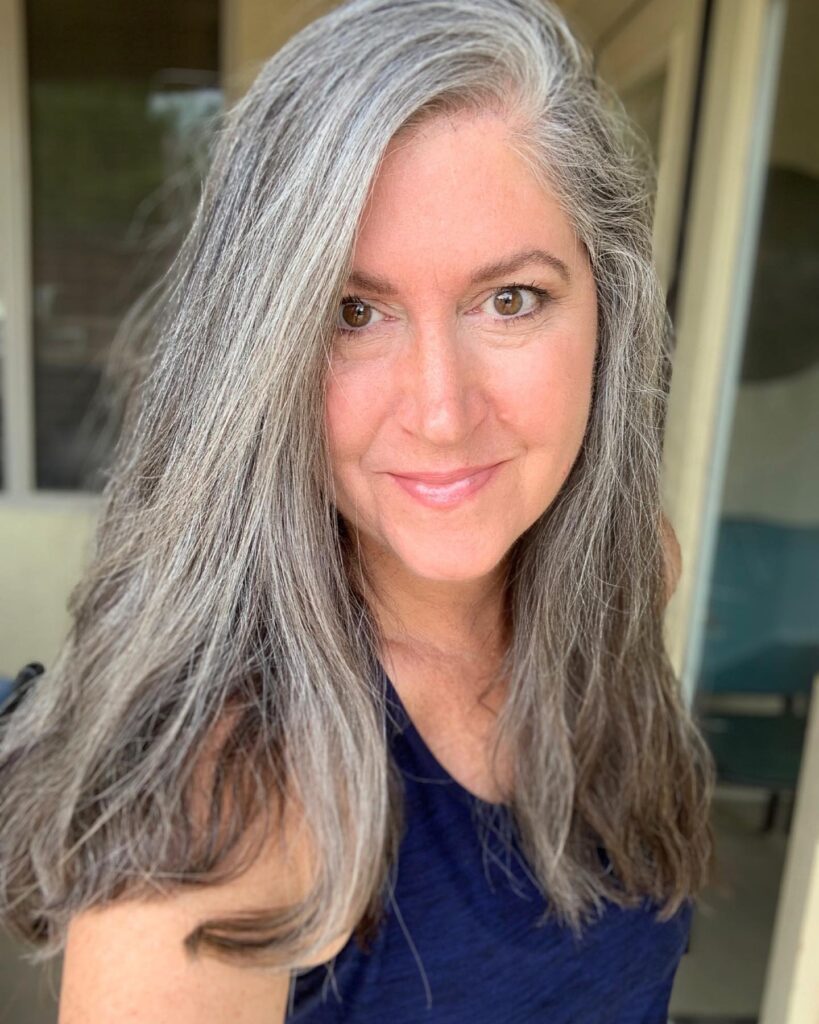 A smiling woman with long salt and pepper hair styled with a casual side flip to reveal bright silver streaks.