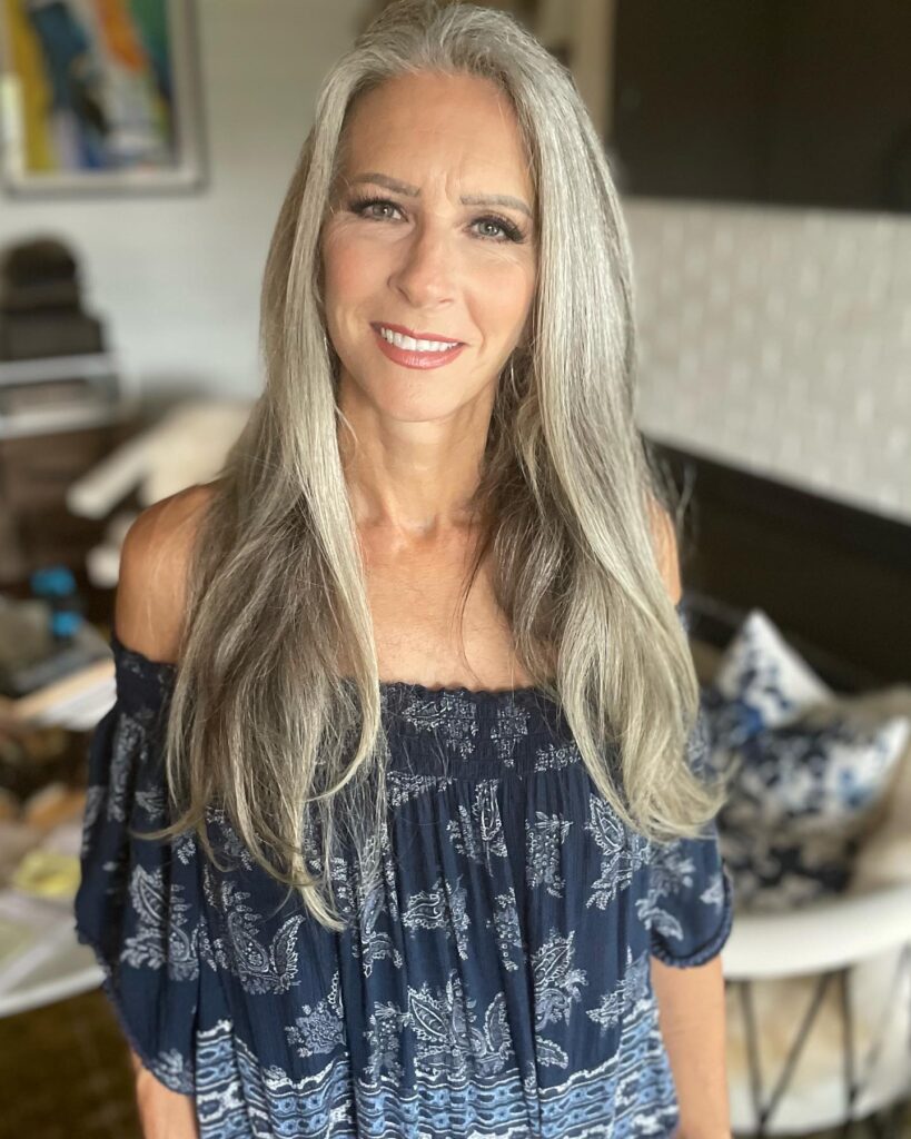 A smiling woman with long straight gray hair wearing a blue off the shoulder patterned top.