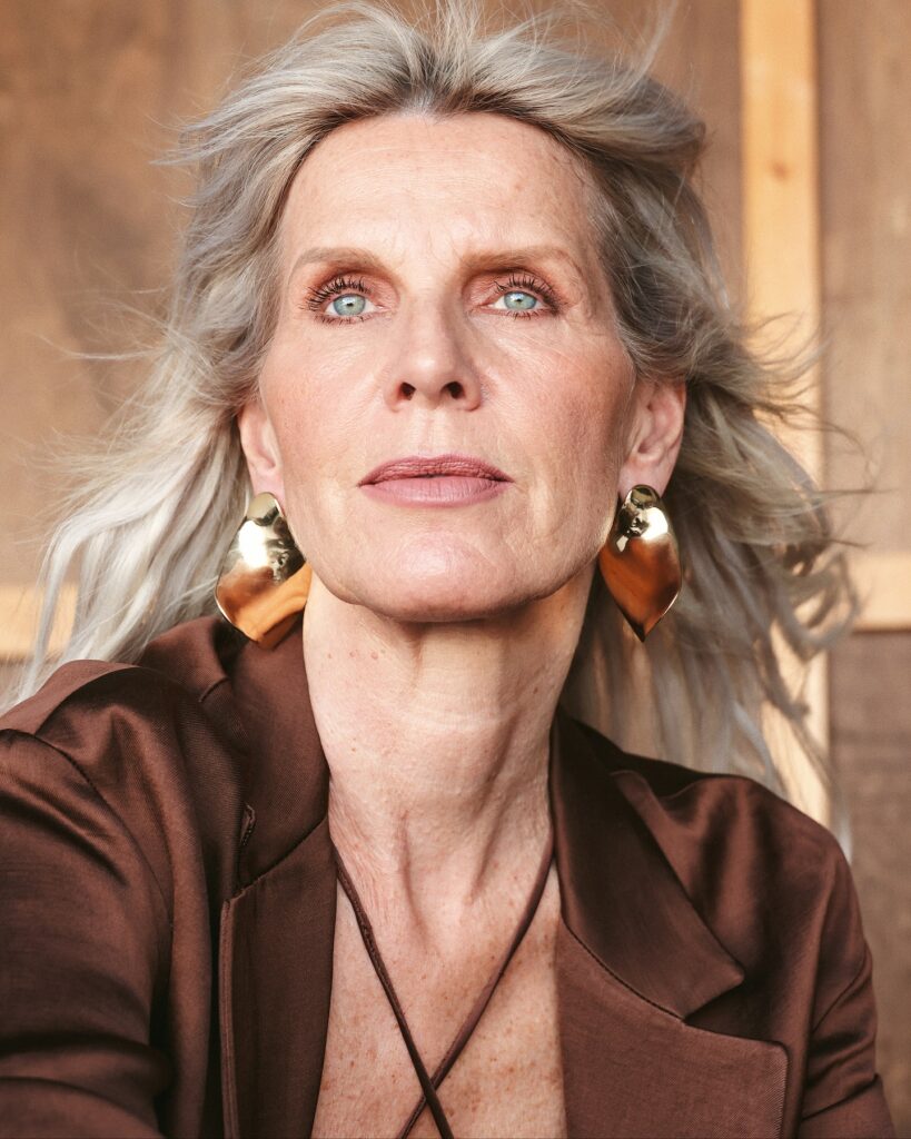 A woman with green eyes and windswept, long platinum silver hair wearing large gold earrings and a brown satin blazer.