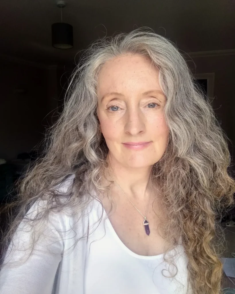 A woman with a purple crystal necklace looks ahead with her long curly silver and golden blonde hair swept softly to the side.