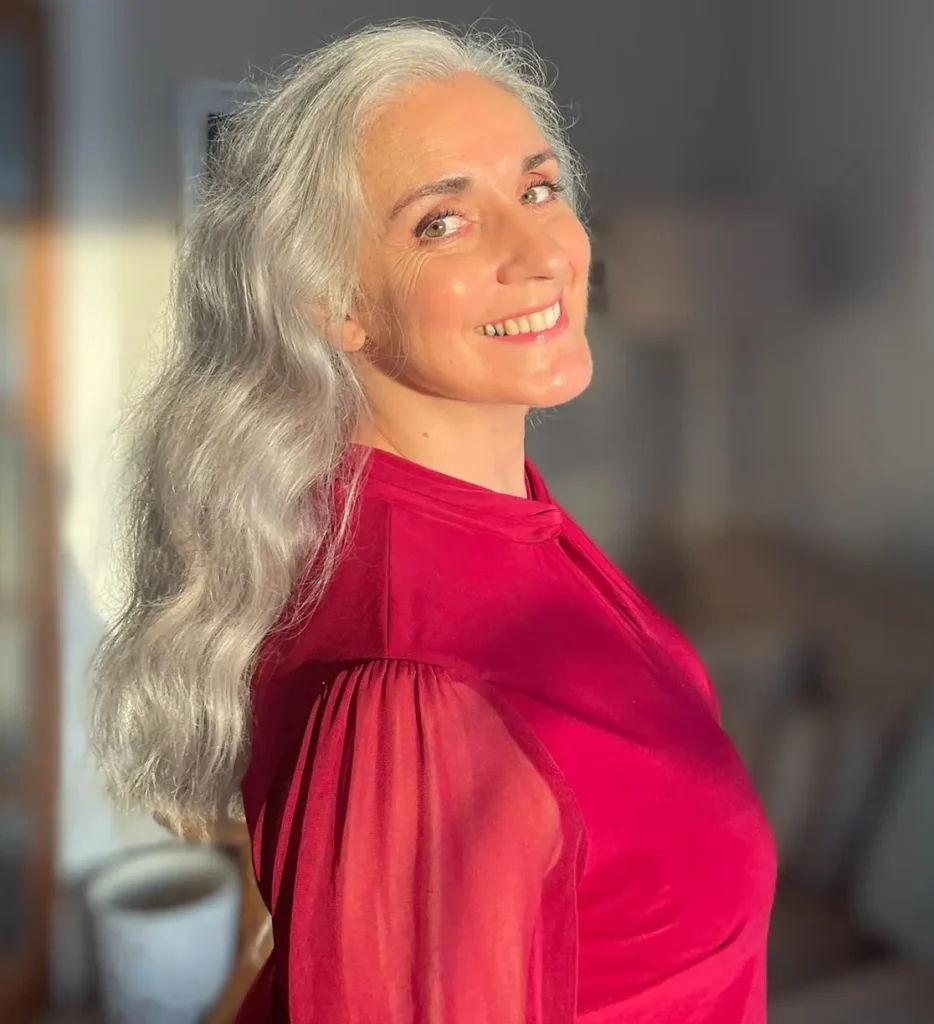 A smiling woman in a vibrant red blouse turns her head to show off her long wavy silver hair catching the warm sunlight.