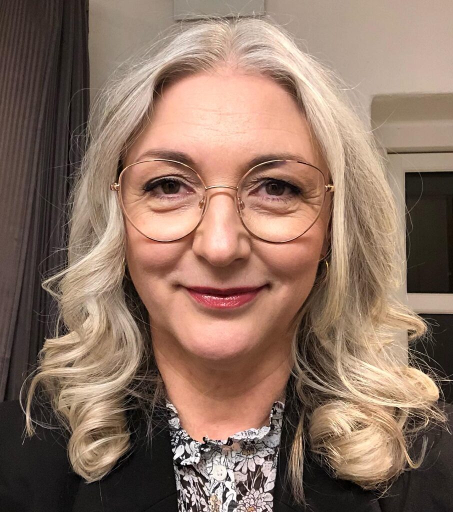 A woman with round glasses wearing a black blazer smiles with her shoulder-length platinum and silver hair styled into defined bouncy ringlets.