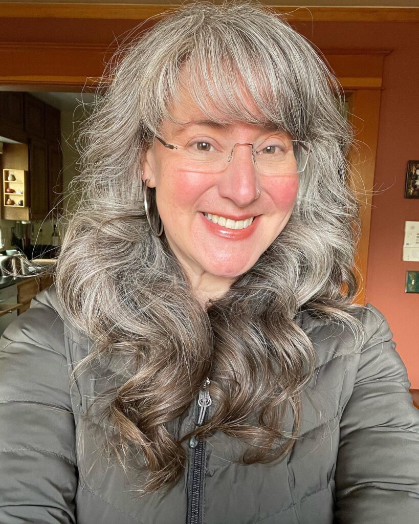 A smiling woman with glasses wearing a green puffer jacket showcases her wavy salt and pepper long hair with wispy bangs.