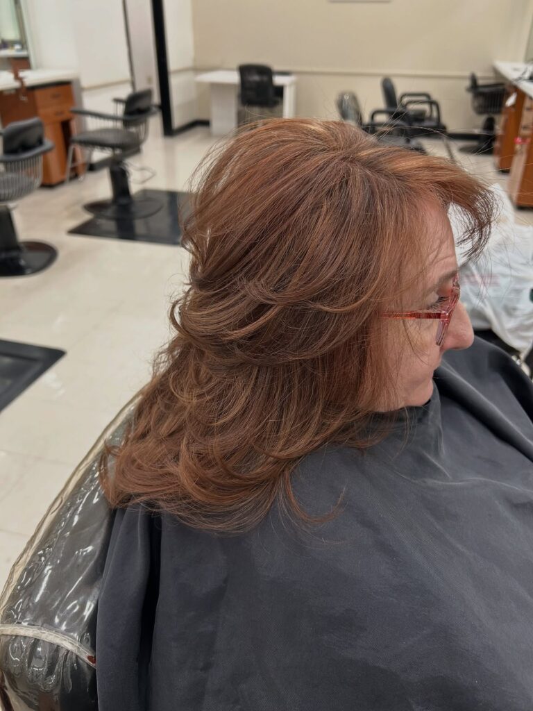 A woman over 60 inside a salon showing the side profile of a layered auburn blowout hairstyle with soft flipped ends.
