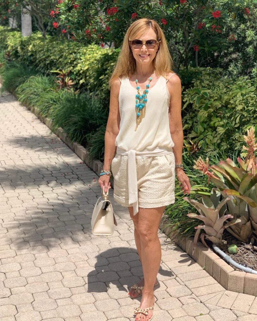 A woman with long voluminous caramel blonde hair and side-swept bangs wearing sunglasses and an cream outfit on a paved pathway.