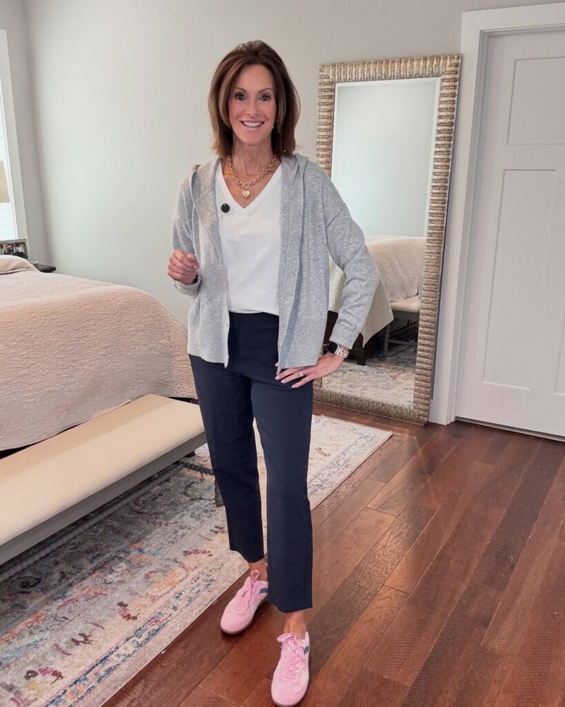 A woman with a voluminous layered chestnut brown lob haircut wearing a grey cardigan and pink sneakers in a bedroom.
