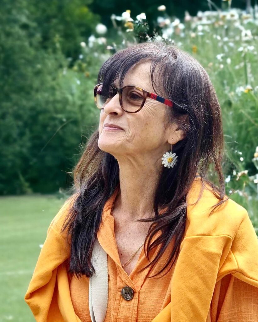 A woman with long layered dark brown hair and wispy side bangs wearing glasses and a bright yellow top in a garden.