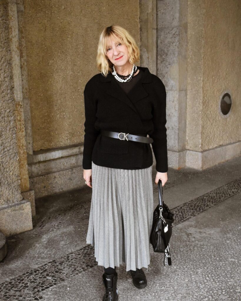 A woman with a chin-length wavy blonde bob haircut and wispy bangs wearing a black jacket and pleated skirt.