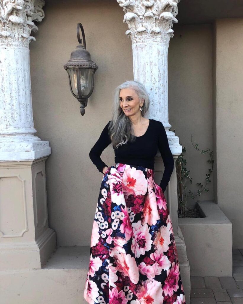 A woman with long cascading silver-gray hair styled with a side part wearing a black top and a large floral print skirt.
