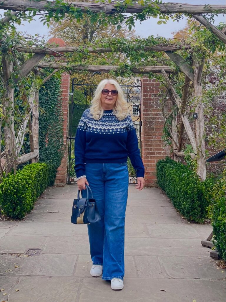 A woman with long, voluminous platinum blonde wavy hair wearing sunglasses and a blue patterned sweater walking under a garden arbor.