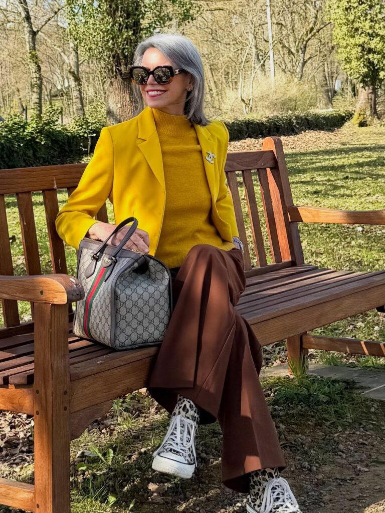 A woman with a straight shoulder-length silver-gray bob haircut sitting on a wooden bench outdoors wearing a bright yellow blazer and sunglasses.