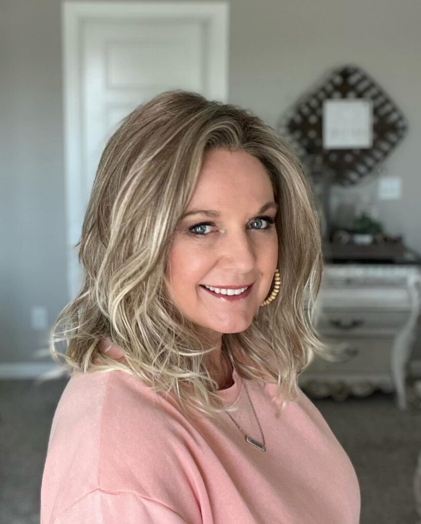 A smiling woman with shoulder-length, voluminous caramel blonde hair styled in deep Hollywood waves wearing a black top and a silver necklace.