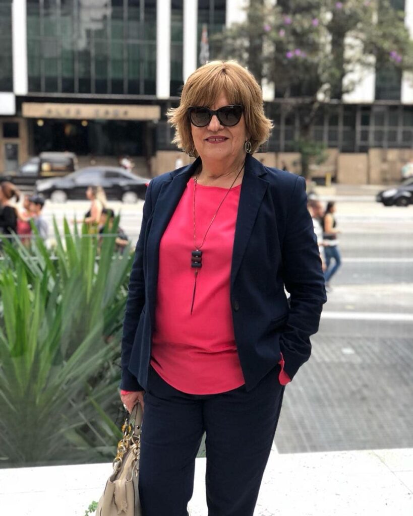 A woman with a short layered golden brown shag haircut and full bangs wearing sunglasses and a navy blazer.