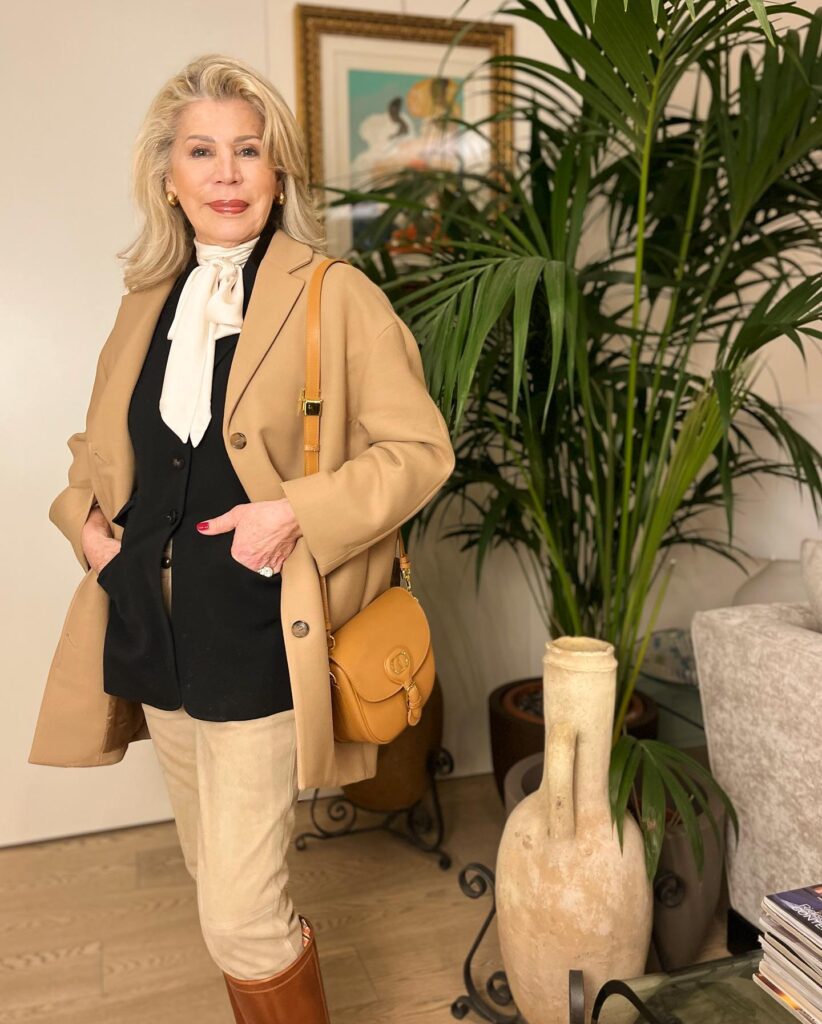 A woman with a voluminous medium-length blonde blowout and swept-back layers styling a camel coat indoors.