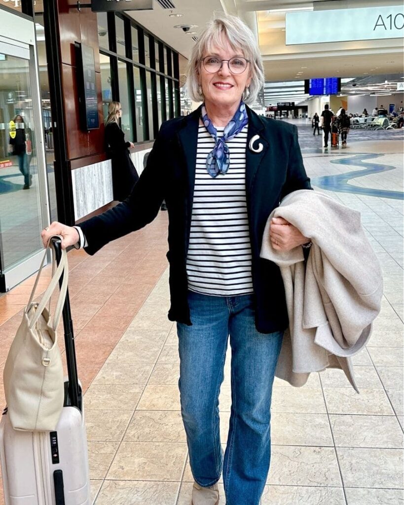 A smiling woman with glasses wearing a striped shirt and blazer showing a neck-length layered ash blonde hairstyle with bangs at an airport.