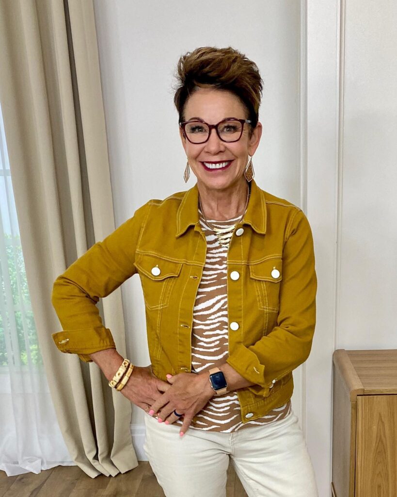 A smiling woman with a short layered silver white pixie bob hairstyle posing indoors in a patterned blouse.
