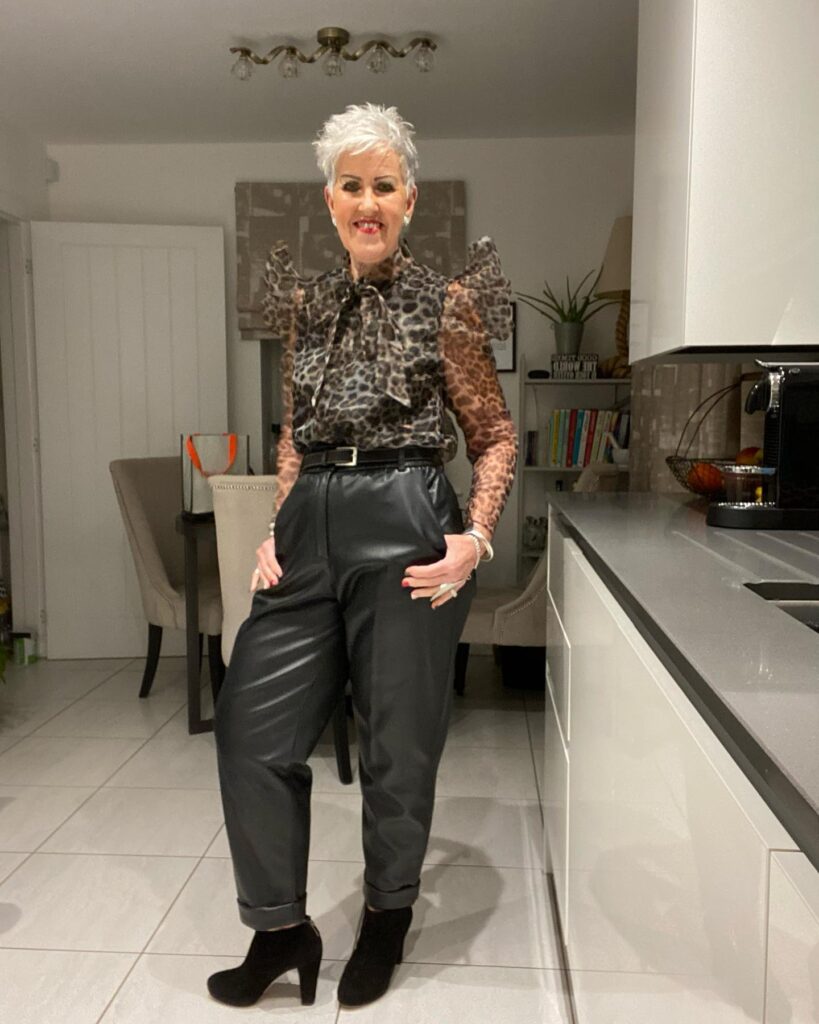 A woman with a short textured platinum white pixie cut posing indoors in a leopard print blouse and black pants.