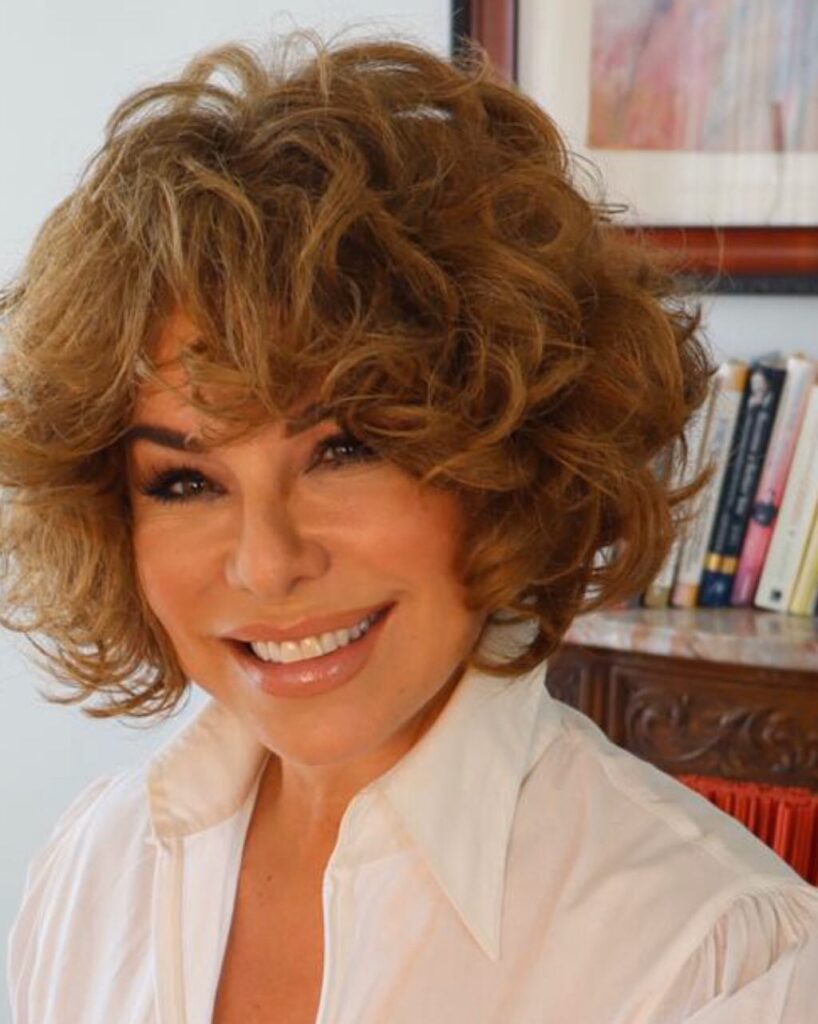 A woman with a voluminous chin-length curly brown shag hairstyle and full bangs smiling.