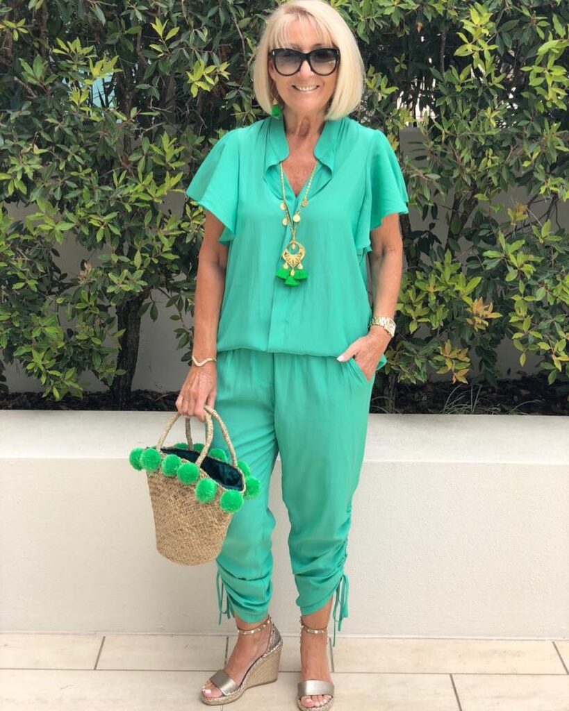 A smiling woman with a smooth chin-length blonde bob and light bangs posing outdoors in a bright green jumpsuit.