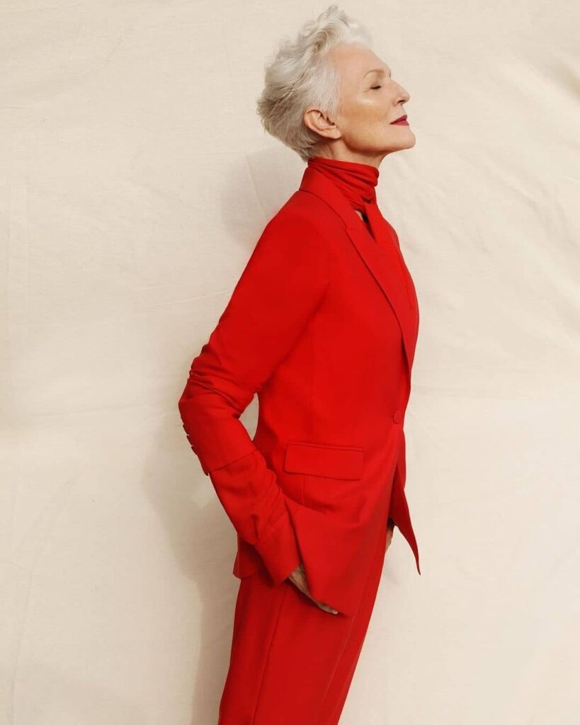 A woman in profile with a short sculpted white pixie cut styled upward wearing a bright red pantsuit.