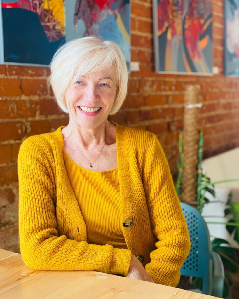 A smiling woman with a short platinum blonde asymmetrical bob hairstyle sitting indoors in a yellow cardigan.