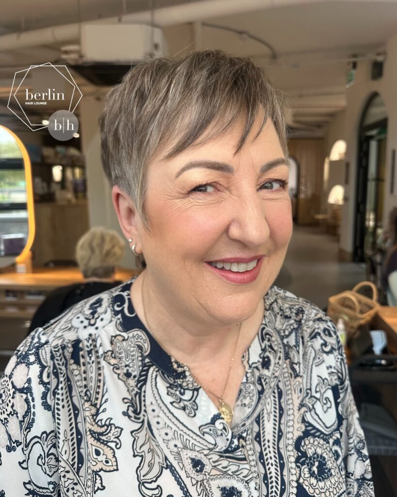 A close-up front view of a smiling woman with a short, wispy salt-and-pepper pixie haircut and soft bangs wearing a paisley-patterned blouse.
