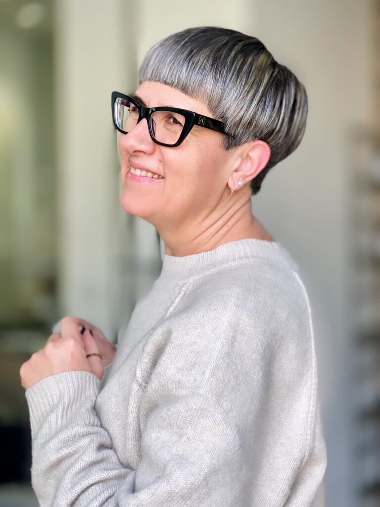 A side profile view of a smiling woman with a geometric silver and charcoal grey bowl-style pixie cut wearing a white sweater and black glasses.