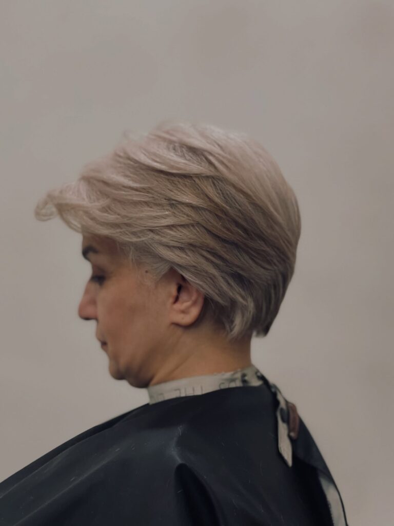 A side profile view of a woman with a feathered, swept-back ash-blonde pixie bob haircut wearing a black salon cape.