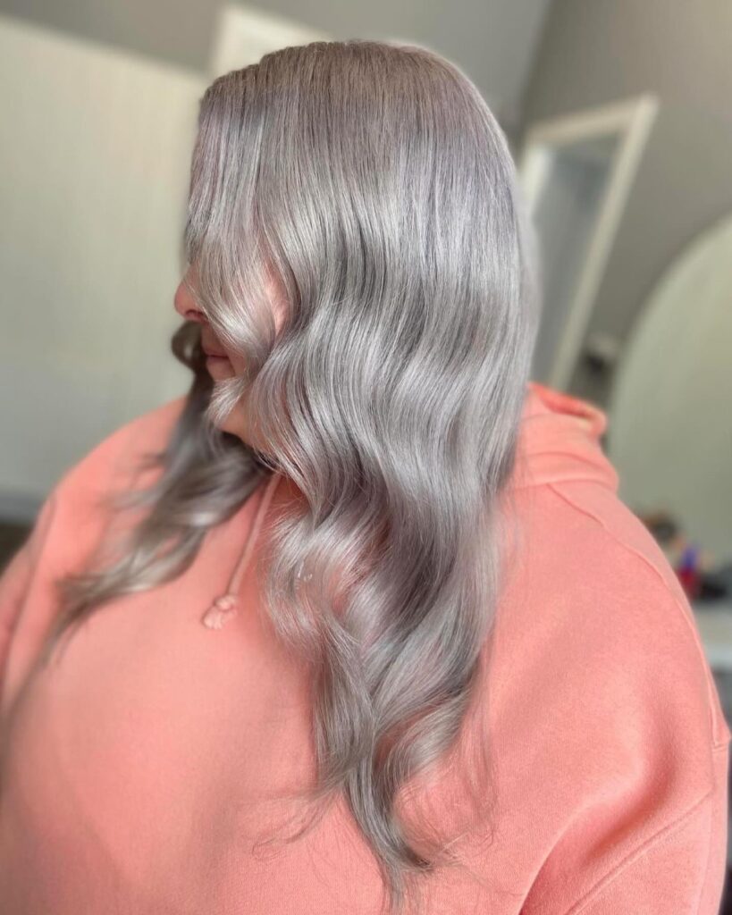 A side-angle view of a woman showcasing long, glossy metallic silver hair styled into soft Hollywood waves while wearing a coral-pink hoodie.