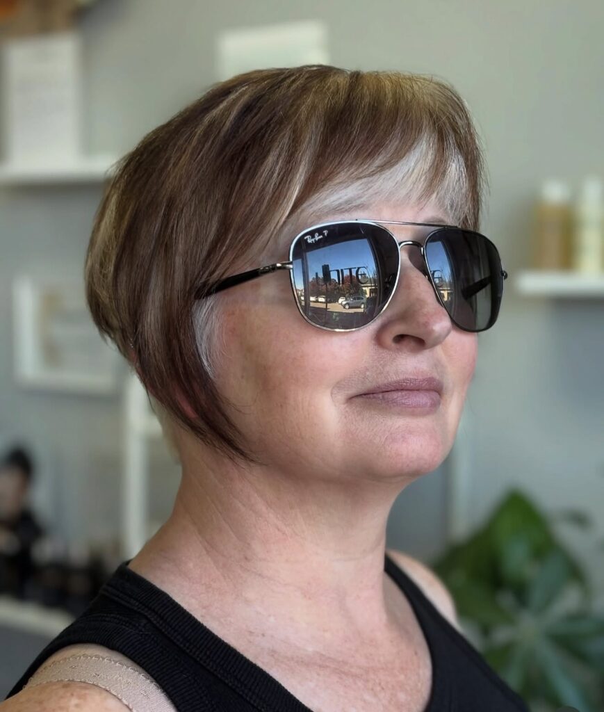 A side profile view of a woman wearing aviator sunglasses and showcasing a two-toned brunette and silver pixie bob haircut.
